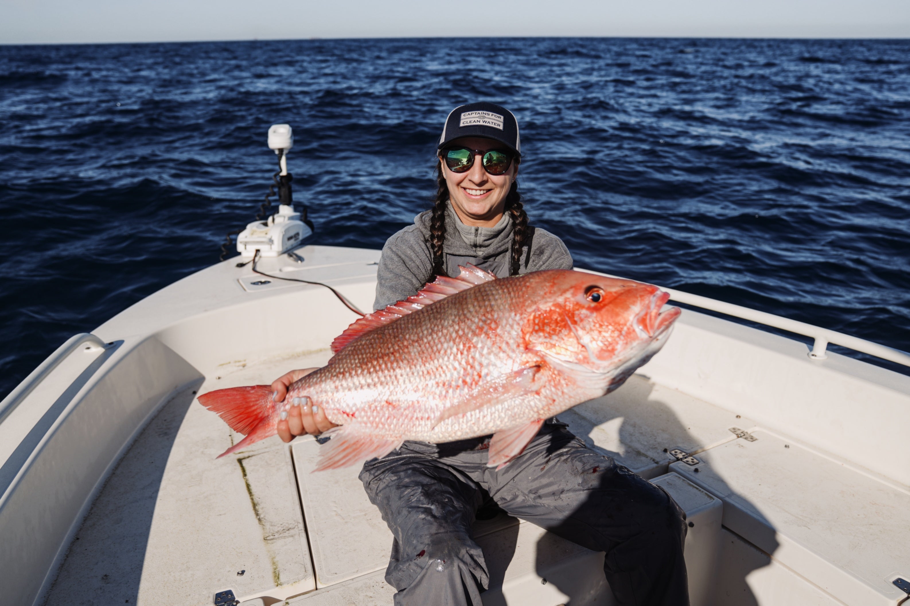 A female Angler is holding a giant Red Snapper while fishing offshore on a boat. She is wearing a Captains for Clean Water hat
