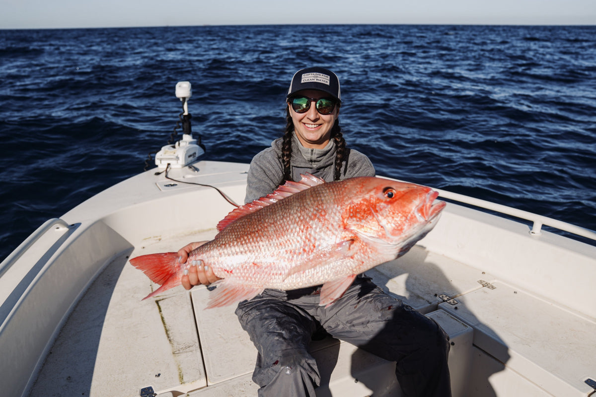 A female Angler is holding a giant Red Snapper while fishing offshore on a boat. She is wearing a Captains for Clean Water hat