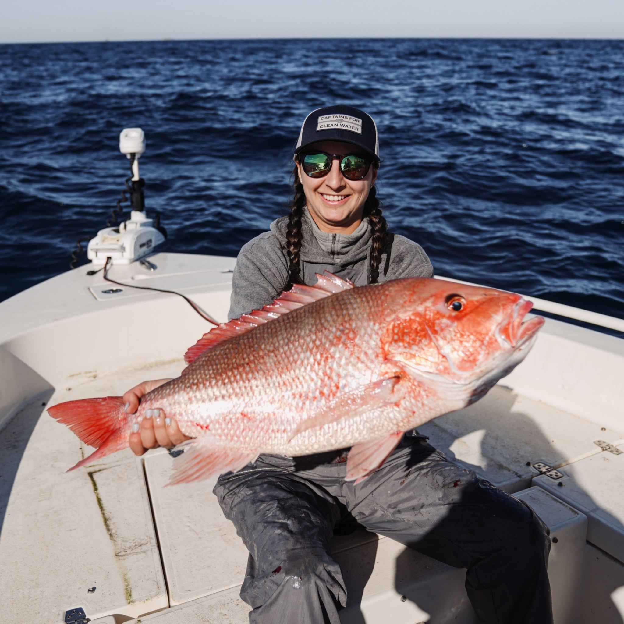 A female Angler is holding a giant Red Snapper while fishing offshore on a boat. She is wearing a Captains for Clean Water hat