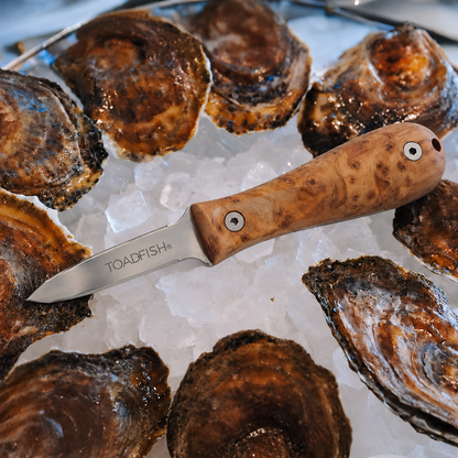 Toadfish Burl Wood Oyster Knife with Pro blade lying diagonally across crushed ice surrounded by oysters